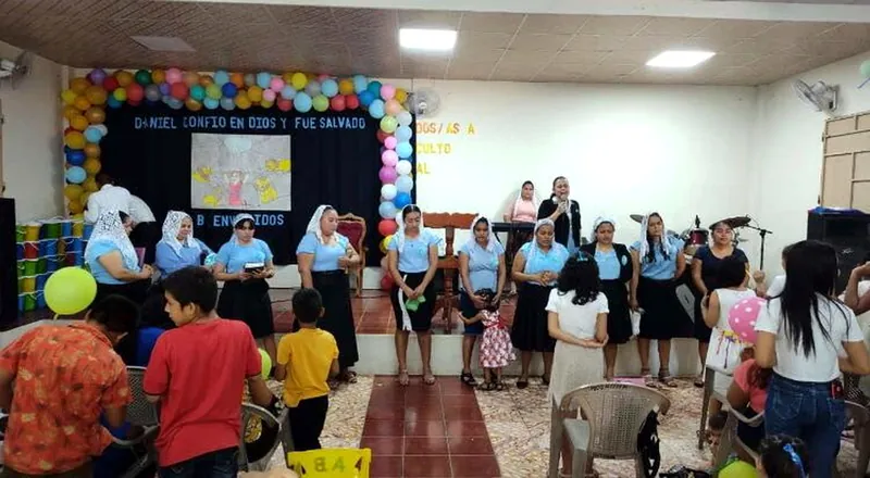 Niños y voluntarios reunidos en un salón comunitario decorado con globos y un cartel.